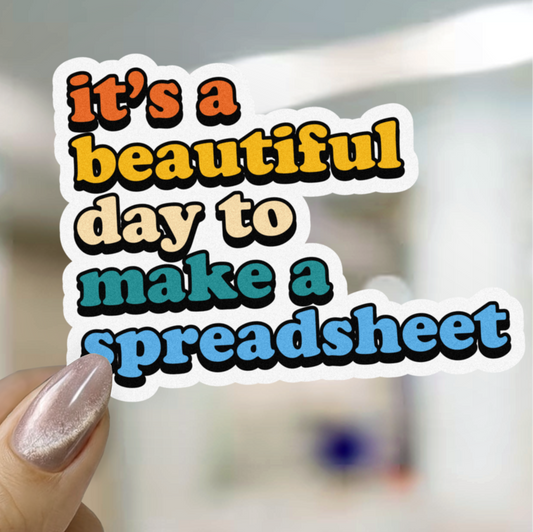 Hand holding a sticker with text 'it's a beautiful day to make a spreadsheet' against a blurred indoor background.