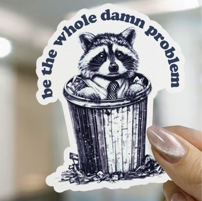 Sticker with a raccoon in a trash can and text 'be the whole damn problem' held by a hand.
